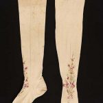 Pair of cream colored knitted stockings with spray of flowers embroidered with polychrome silks on instep; stained.